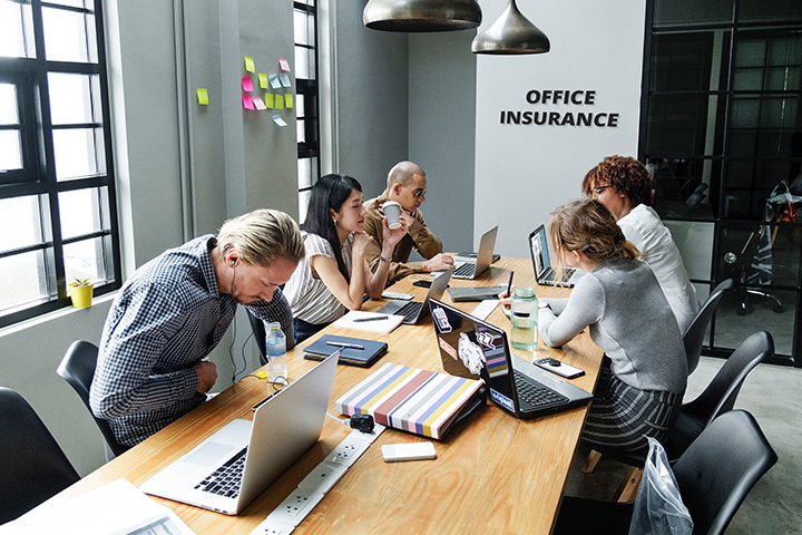 A group of people in a board room in an office building, they all have laptops and notebooks and on the wall behind them are the words office insurance