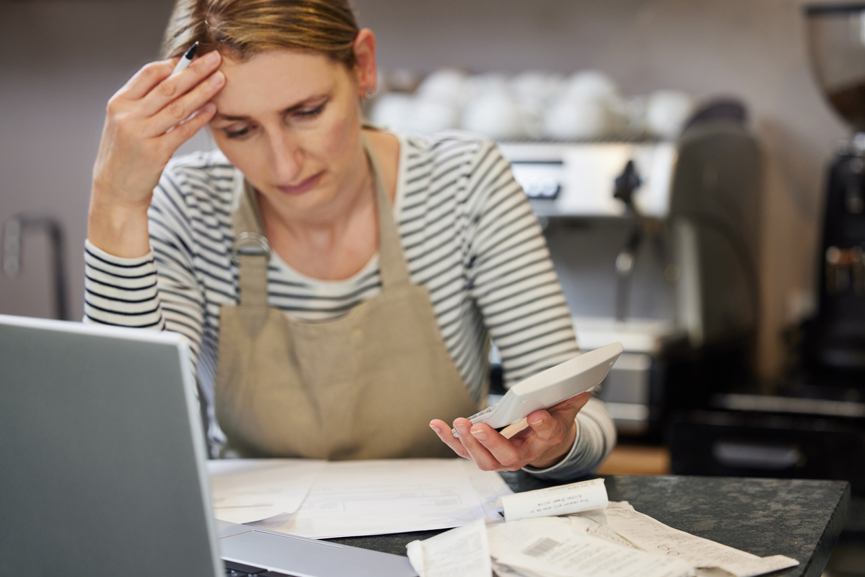 Worried Female Owner Of Coffee Shop In Financial Difficulty Looking Through Bills Using Laptop And Calculator