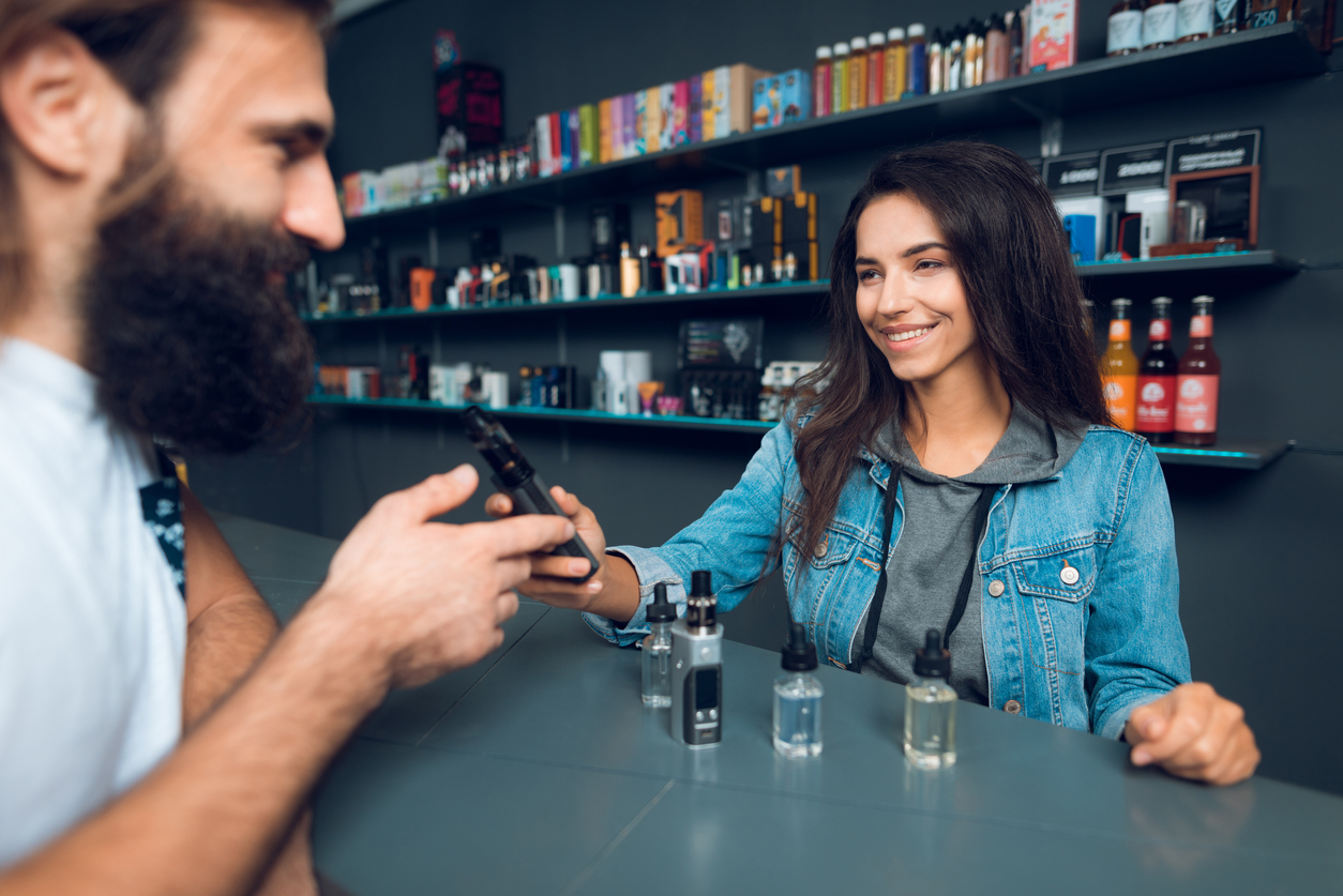 Girl seller shows the choice of electronic cigarettes in vapeshop. Nearby is a buyer - a man with a beard. The store has a large assortment of electronic cigarettes.