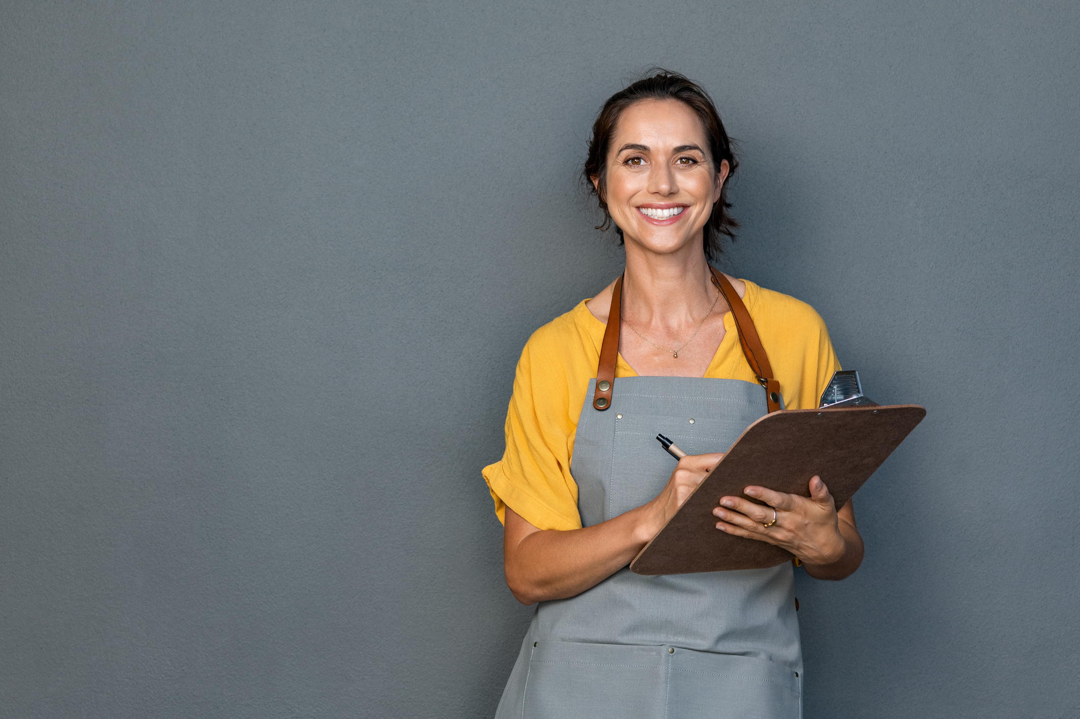 Happy smiling waitress taking orders isolated on grey wall. Mature woman wearing apron while writing on clipboard standing against gray background with copy space. Cheerful owner ready to take customer order while looking at camera. Small business concept.