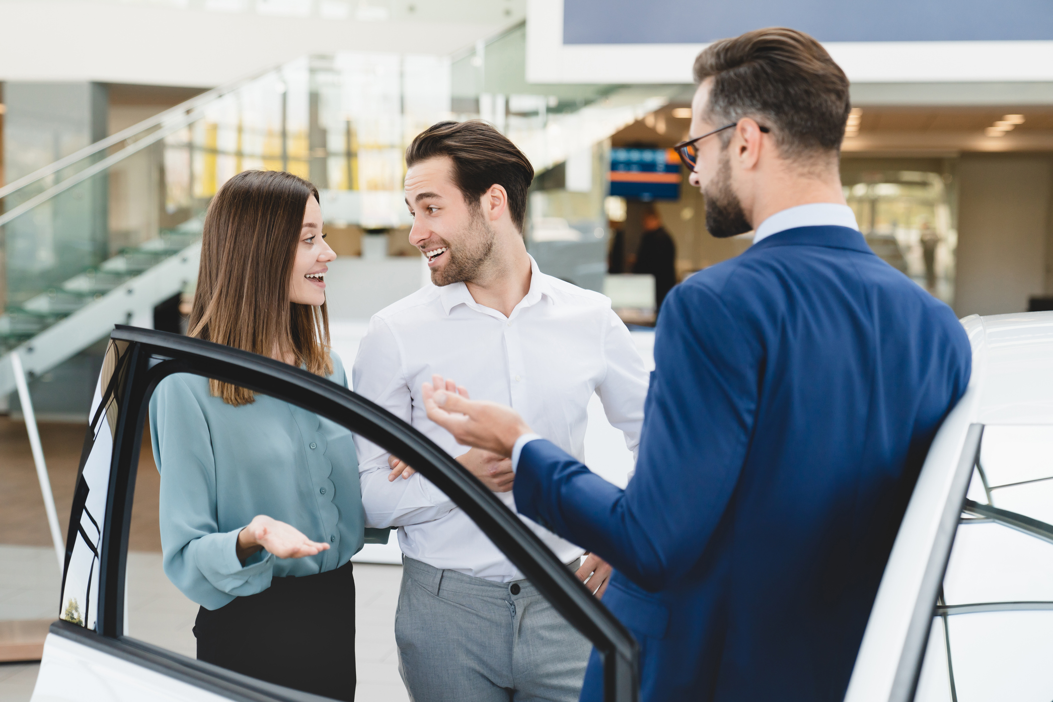 Car test-drive concept. Male shop assistant in formal clothes helping caucasian young family in choosing buying purchasing new car at automobile dealer shop store.